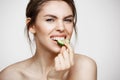 Pretty young natural girl with perfect clean skin looking at camera eating cucumber slice over white background. Facial