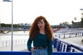 Pretty young red-haired woman is leaning on a railing in a park. She is posing for the photo smiling. Fashion and beauty concept