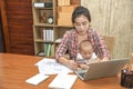 Pretty young single mom working at home on a laptop computer while holding her baby girl sitting on her lap enjoying watching Royalty Free Stock Photo