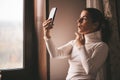Pretty young smiled Caucasian woman in white turtleneck smiling and videochatting on mobile phone in home.Girl talking Royalty Free Stock Photo