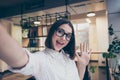 Pretty young smiling girl in glasses taking a selfie working in light room workplace workstation grimacing saying hello hi to her