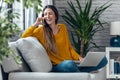 Pretty young smiling woman talking with mobile phone while working with laptop sitting on a couch at home Royalty Free Stock Photo