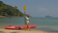 Pretty young sportswoman with sunglasses and swimsuit holds paddle posing near pink plastic canoe on sand beach at Royalty Free Stock Photo