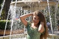 Pretty young woman, blonde, blue eyes, in a green flowered suit with her hands in her hair, smiling, with a water fountain behind