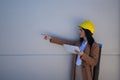 Pretty young woman in brown coat, yellow workman`s helmet and blueprint tube, working with her tablet and pointing with her hand. Royalty Free Stock Photo