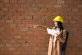 Pretty young woman in brown coat, yellow workman`s helmet and blueprint tube, working with her tablet and pointing with her hand. Royalty Free Stock Photo