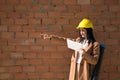 Pretty young woman in brown coat, yellow workman`s helmet and blueprint tube, working with her tablet and pointing with her hand. Royalty Free Stock Photo