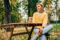 Pretty young woman drinking coffee at the terrace cafeteria in the park. Blonde female in the city street drinking a hot beverage