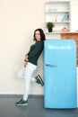 Pretty young woman in a green sweater and light jeans and sneakers stands leaning on a blue retro refrigerator in the kitchen in