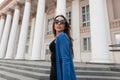 Pretty young woman hipster in sunglasses in a stylish blue knitted cape in a stylish black top with lace in jeans poses Royalty Free Stock Photo