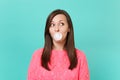 Pretty young woman in knitted pink sweater looking up, chewing, blowing bubble gum balloon isolated on blue turquoise Royalty Free Stock Photo