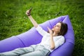 Pretty young woman lying on inflatable sofa lamzac send kisses on video call or take selfie while resting on grass in park on the Royalty Free Stock Photo