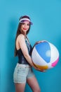 Pretty young woman posing in swimsuit and pink plastic visor cap with inflatable ball on blue background Royalty Free Stock Photo