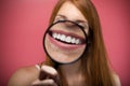 Pretty young woman showing teeth trought magnifying glass over pink background.