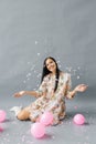 A pretty young woman sits near pink balloons and throws up confetti and smiles and on a gray background Royalty Free Stock Photo