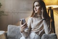 Pretty young woman sitting on sofa and listening to music on smartphone. Girl in earphones streaming songs online in social