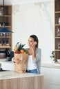 Pretty young woman talking on her mobile phone while holding shopping bag with fresh vegetables in the kitchen Royalty Free Stock Photo