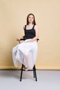 Pretty young woman in tank top and skirt sitting on chair over pale yellow background