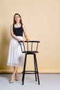 Pretty young woman in tank top and skirt standing near bar chair over pale yellow background