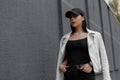 Pretty young woman in a trendy T-shirt in a white stylish leather jacket in a black fashionable cap poses near a vintage gray wall Royalty Free Stock Photo