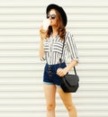Pretty young woman walking with coffee cup in black round hat, shorts, white striped shirt on white wall