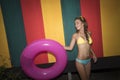 Pretty young woman wearing bikini holding pink inflatable ring on the colourful wall