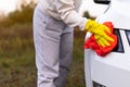 A pretty young woman in a white hoodie and yellow rubber gloves wipes the headlights of a car with a bright red rag on a warm autu Royalty Free Stock Photo