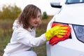 A pretty young woman in a white hoodie and yellow rubber gloves wipes the headlights of a car with a bright red rag on a warm autu Royalty Free Stock Photo