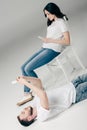 Young woman sitting on chair and using smartphone near handsome man lying on floor and taking selfie on grey background Royalty Free Stock Photo