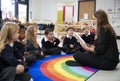 Primary school kids sitting on the floor in class listening to their female teacher reading a book to them, side view