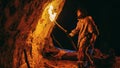 Primeval Caveman Wearing Animal Skin Exploring Cave At Night, Holding Torch with Fire Looking at D
