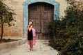 A sweet princess girl in a crown and a burgundy dress with a pink veil is pampered in front of an old house with an ancient wooden