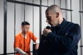 A prison guard in full ammunition communicates with his partner on the radio. In the background, a prisoner in a cell