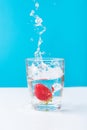 Process of falling of ripe juicy strawberry into glass of fresh pure water with splashes bubbles blue white background. Hydration