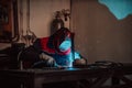Profesional welder in protective uniform and mask welding metal pipe on the industrial table with other workers behind