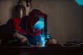 Profesional welder in protective uniform and mask welding metal pipe on the industrial table with other workers behind