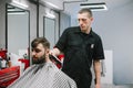 Professional barber cuts the hair of an adult man with a serious face, uses a clipper. Portrait of male hairdresser at work in Royalty Free Stock Photo