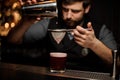 Professional bartender with a beard pouring a smooth dark red cocktail through the sieve to the glass with one big ice