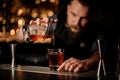 Professional bartender pouring an alcohol from the measuring cup through the strainer to the glass with one big ice cube