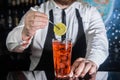 A professional bartender puts a slice of lemon in a red chilled alcoholic cocktail with bar tweezers in glass at a nightclub