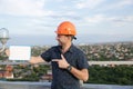 Builder in a protective helmet with a tablet for writing in his hand is standing on the roof of a building overlooking the city Royalty Free Stock Photo