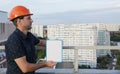Builder in a protective helmet with a tablet for writing in his hand is standing on the roof of a building overlooking the city Royalty Free Stock Photo
