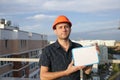 Builder in a protective helmet with a tablet for writing in his hand is standing on the roof of a building overlooking the city Royalty Free Stock Photo