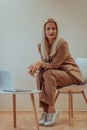 A professional businesswoman sits on a chair, surrounded by a serene beige background, diligently working on her laptop