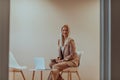 A professional businesswoman sits on a chair, surrounded by a serene beige background, diligently working on her laptop