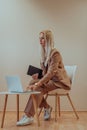 A professional businesswoman sits on a chair, surrounded by a serene beige background, diligently working on her laptop