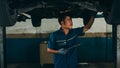 Professional car mechanic using paperwork makes the oil and engine check to the car on lifted automobile at repair service station