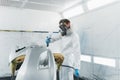 Professional car painter in a protective suit and mask varnishes a painted bumper of a vehicle while working in a painting booth. Royalty Free Stock Photo