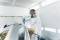 Professional car painter in a protective suit and mask varnishes a painted bumper of a vehicle while working in a painting booth. Royalty Free Stock Photo