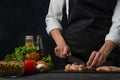 The professional chef in black apron cuts with knife chicken fillet on black chopped board on dark blue background. Backstage of Royalty Free Stock Photo